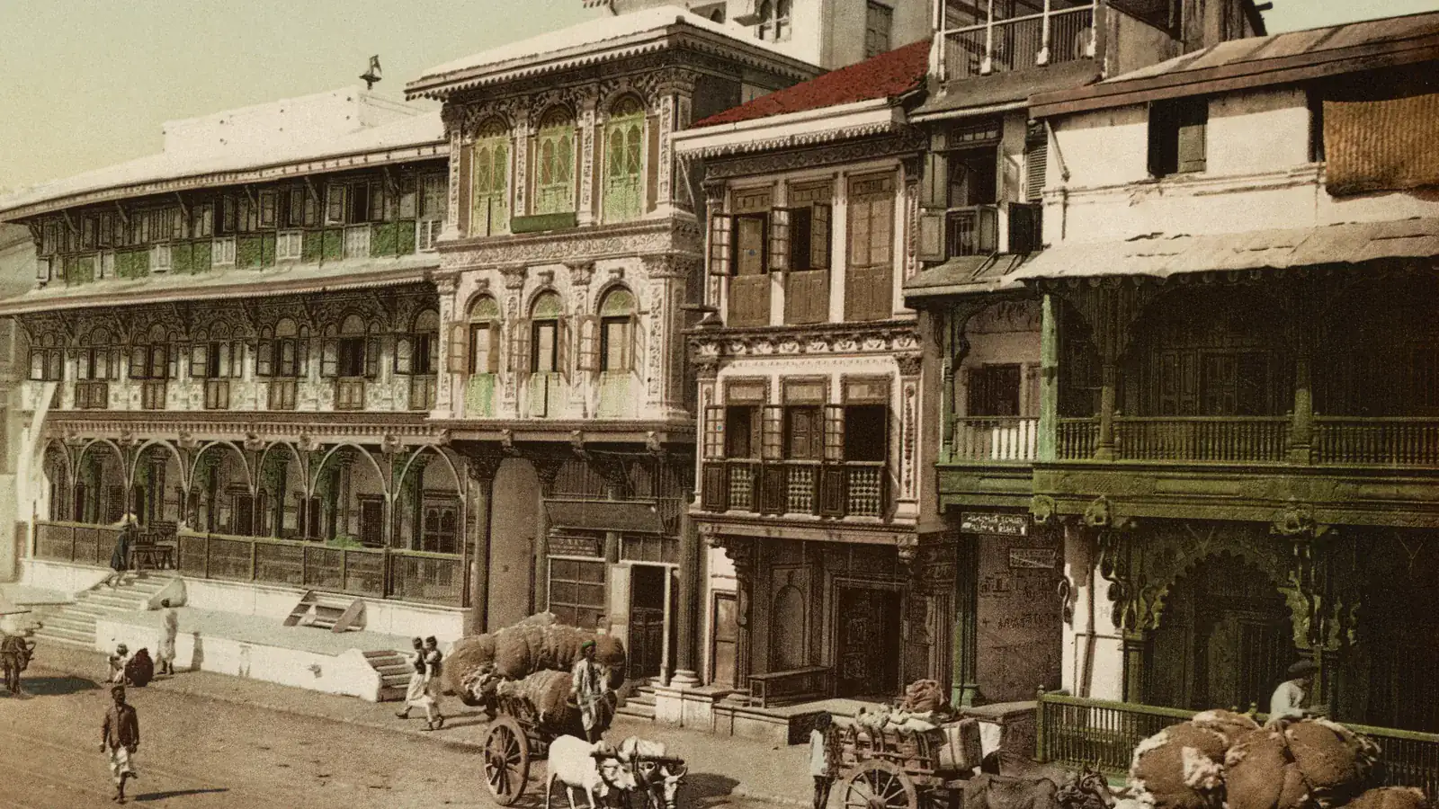 Bombay. Pydownee Street, a colour photochrom of about 1890: carved wooden merchant houses with green shutters above a dusty street where ox carts carry bales