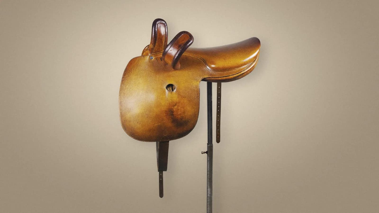 A honey-coloured leather side saddle with two dark leather pommels, made by Hermès in Paris between 1876 and 1925, on its museum stand against a soft warm backdrop