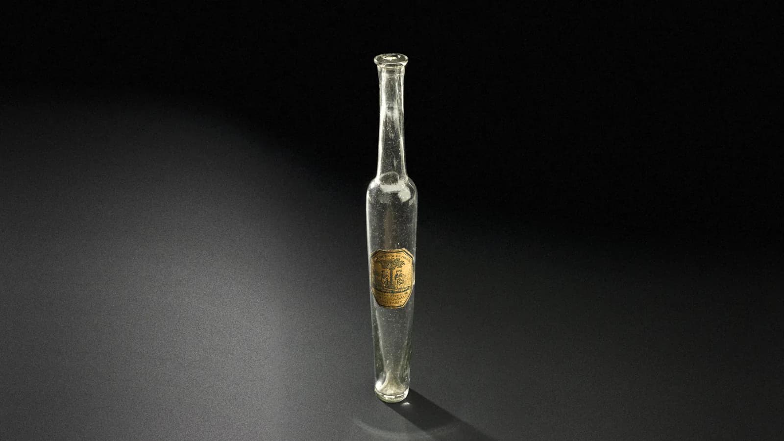 A slim clear glass eau de Cologne bottle made in Paris between 1780 and 1850, with a small engraved paper label, standing on a dark museum backdrop