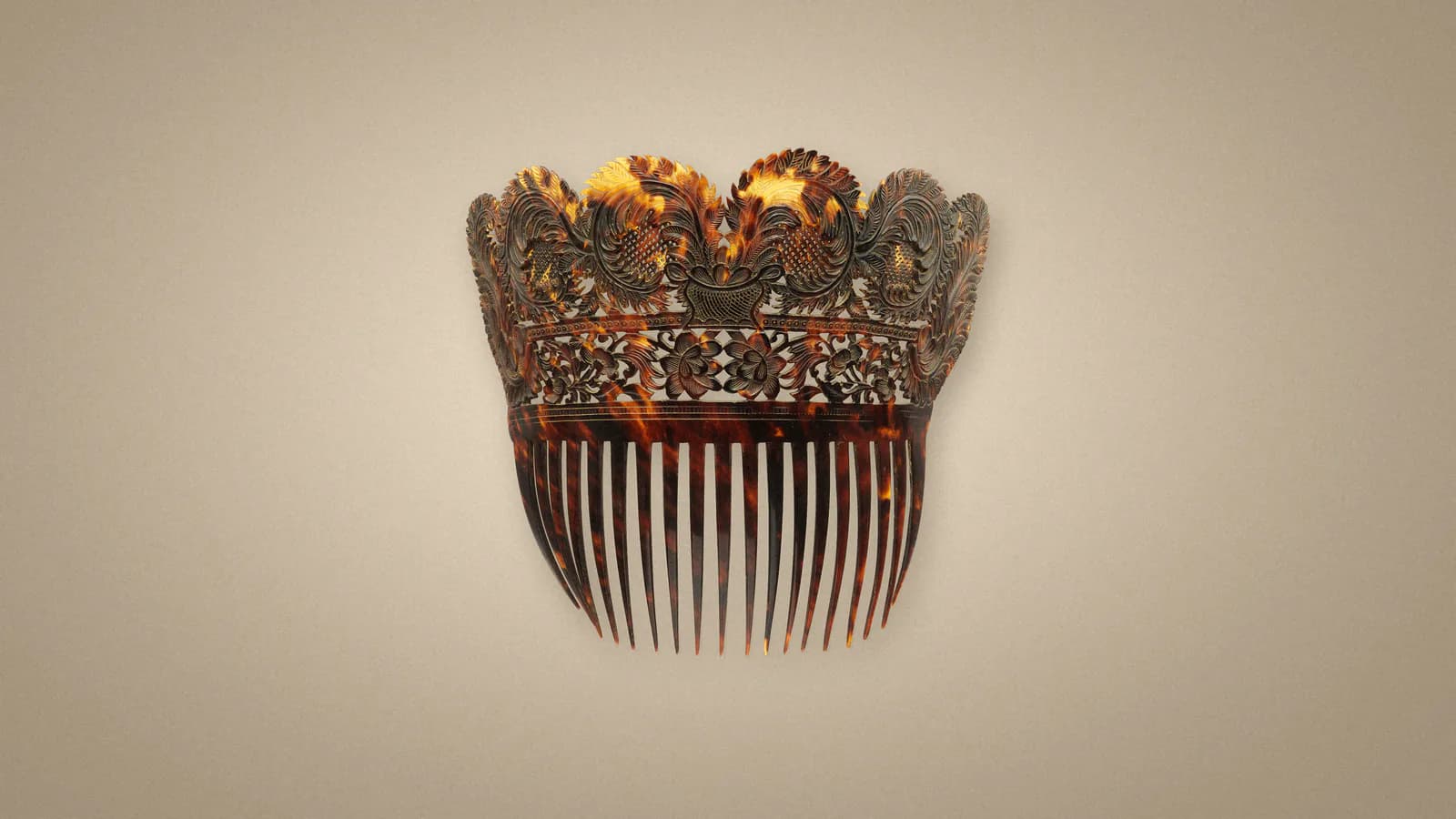 A carved late 18th-century tortoiseshell comb from The Met's open collection, its crown-shaped top of openwork leaves, flowers and a basket glowing amber over long dark teeth, on a soft warm stone-grey ground