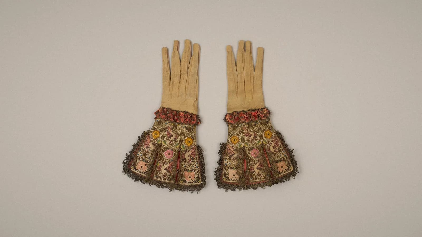 A pair of early 17th-century leather gloves with embroidered satin gauntlets, ribbon ruffs and metal lace, laid flat on a warm grey backdrop