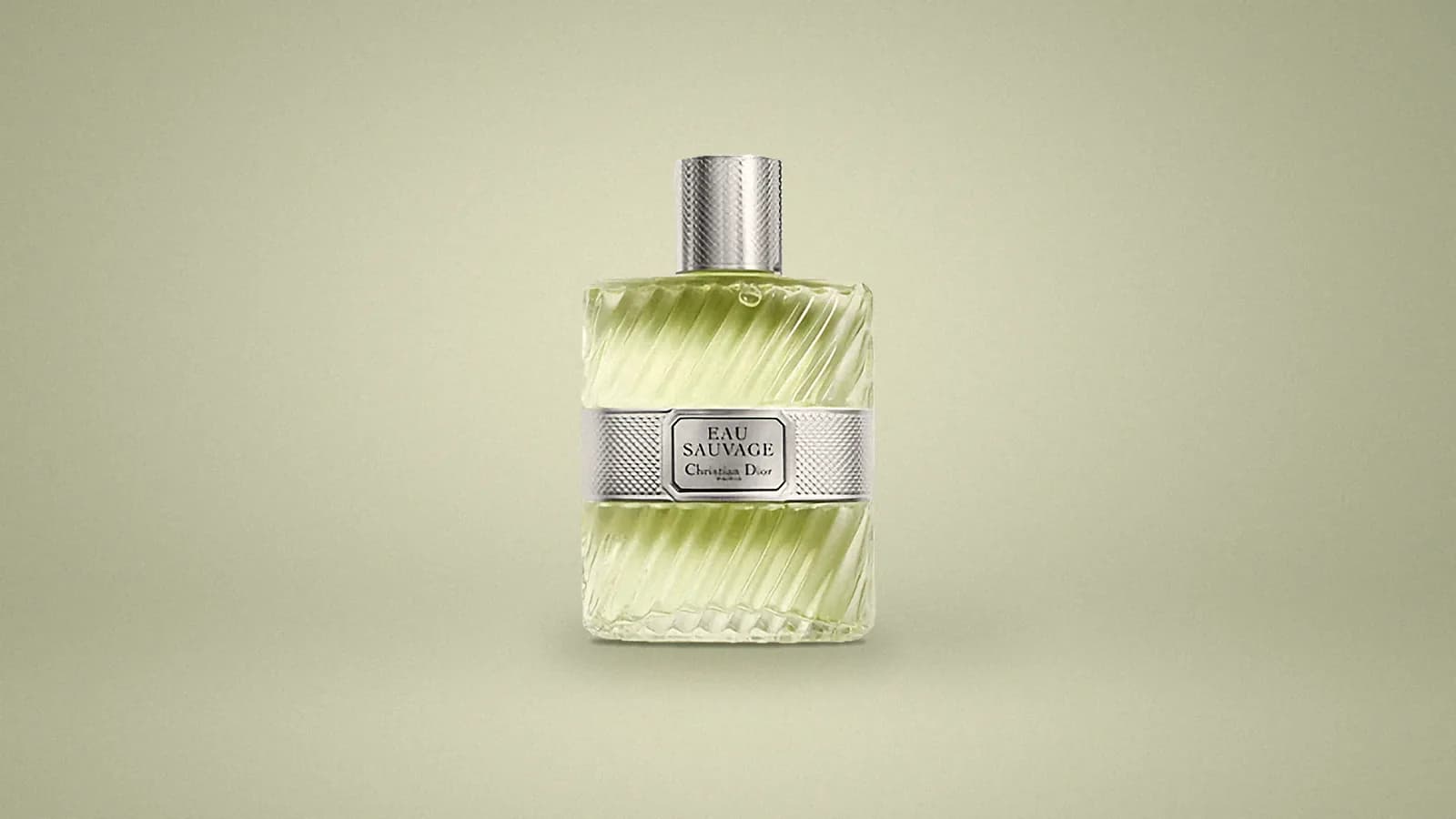Our photo of a Dior Eau Sauvage bottle, a ribbed glass flask of pale green-gold eau de toilette with a knurled silver cap and a silver label band, on a soft sage studio backdrop